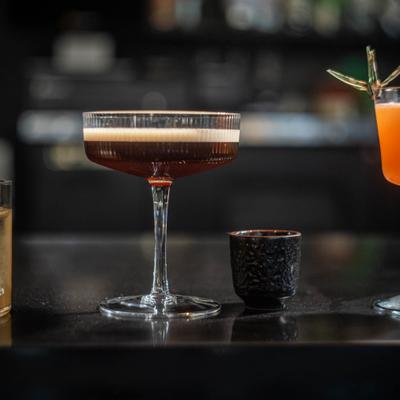 A dark cocktail in a coupe glass sits on a bar counter, with a bright orange drink.