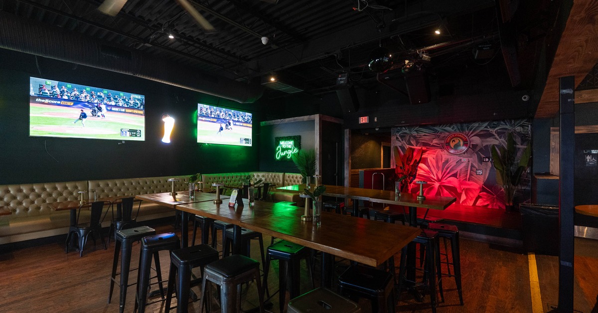 Interior of a bar with two TVs showing a baseball game, a long wooden table with black metal stools