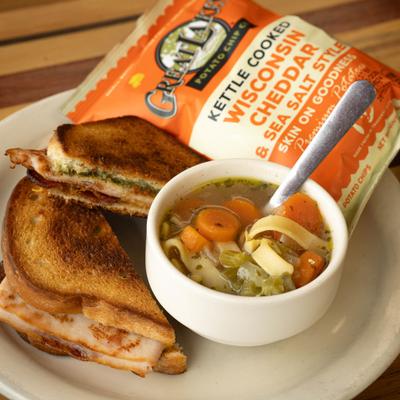 Comforting meal of a toasted sandwich, a bowl of noodle soup, and a bag of chips.