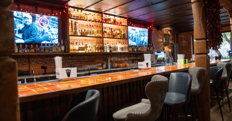A cozy bar interior with a long tiled counter, illuminated shelves with bottles, bar stools