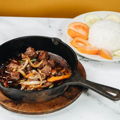 A skillet of Beef Stir Fried, served with a plate of rice, cucumber, and tomato slices.