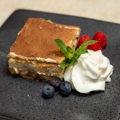 Tiramisu slice served with whipped cream, fresh berries, and mint on dark plate.