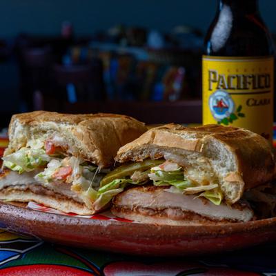 Mexican torta sandwich with chicken, cheese, and lettuce, paired with a beer.