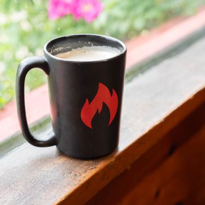 Chai latte in a black mug with a fire logo, sitting on a window.