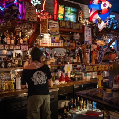 A bartender stands at a dimly lit, lively bar with shelves full of bottles and neon signs.