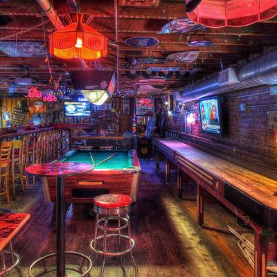Dimly lit bar with a pool table, bar stools, and wooden floors, neon lights and colorful decor
