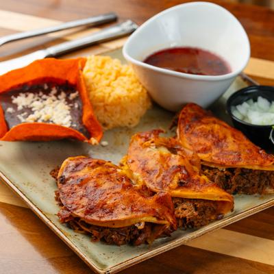 Quesabirria tacos served with dipping broth, refried beans, rice, and chopped onions.