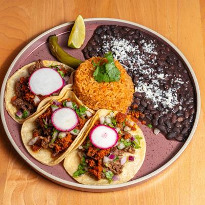 A plate with three tacos served with Mexican rice, black beans, a lime wedge.