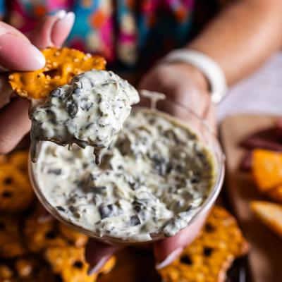 A hand scooping spinach dip with a pretzel chip.