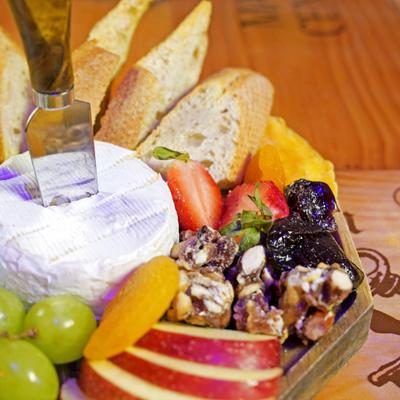 A cheese platter featuring a wheel of Brie cheese, fresh fruits, bread, nuts.