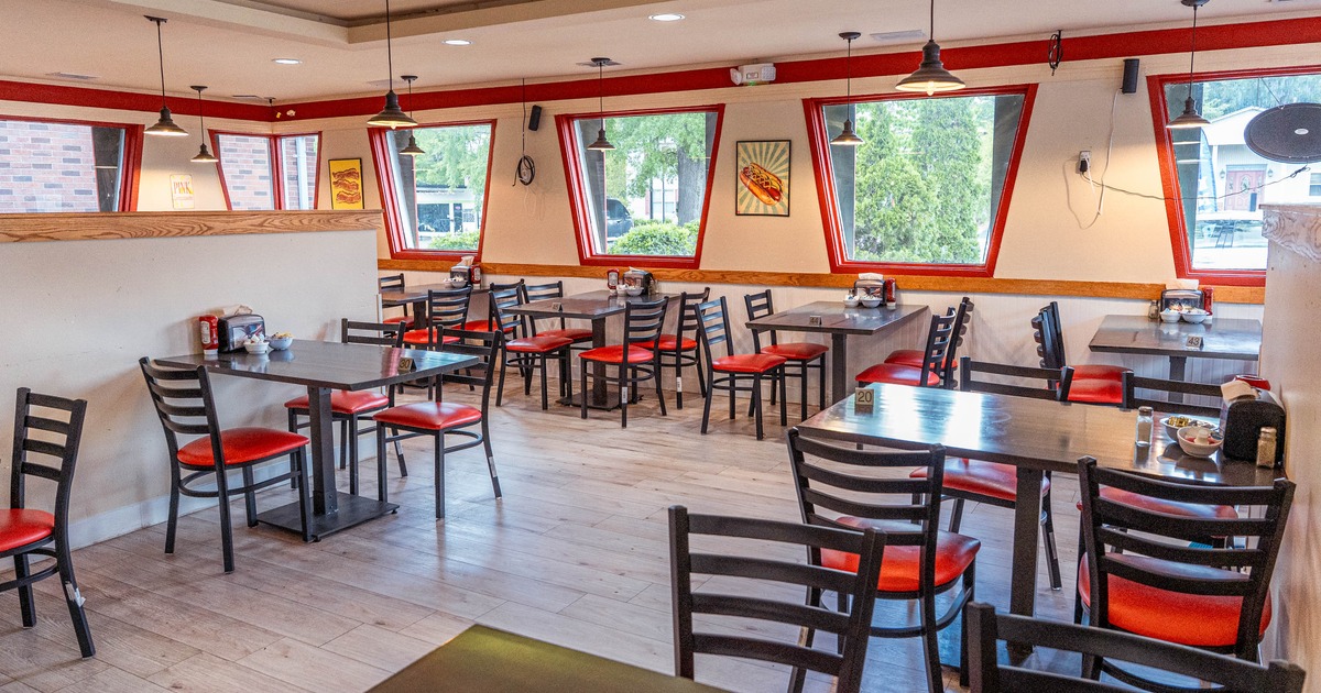 Interior of a restaurant with wooden tables and chairs, red accents and large windows.