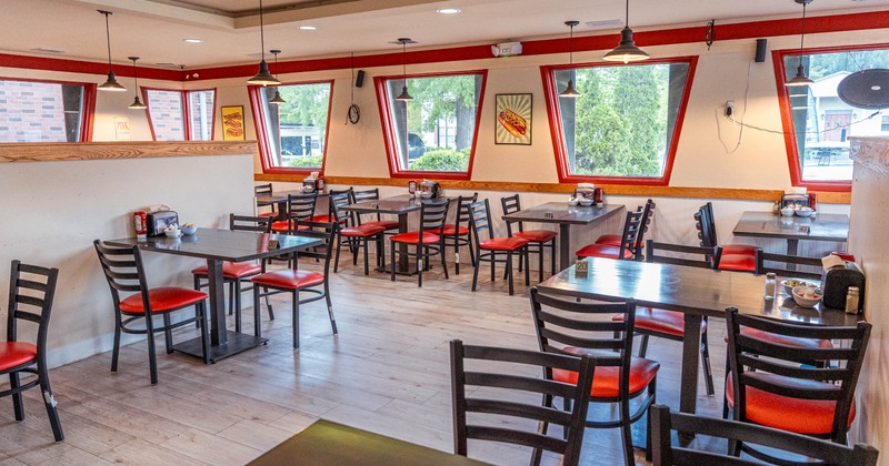 Interior of a restaurant with wooden tables and chairs, red accents and large windows.