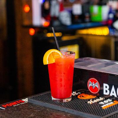 A refreshing red cocktail garnished with an orange slice and a straw, sits on a bar counter