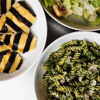 Striped ravioli, pesto fusilli, and Caesar salad in white bowls.