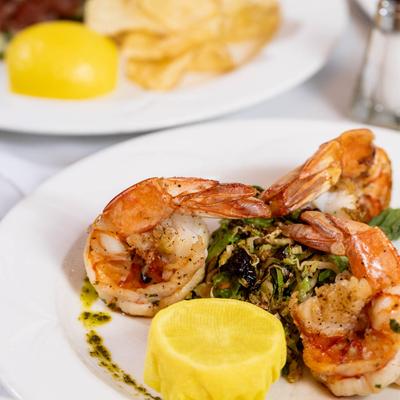 Grilled shrimp atop sauteed greens on a white plate with green sauce drizzle and lemon half.