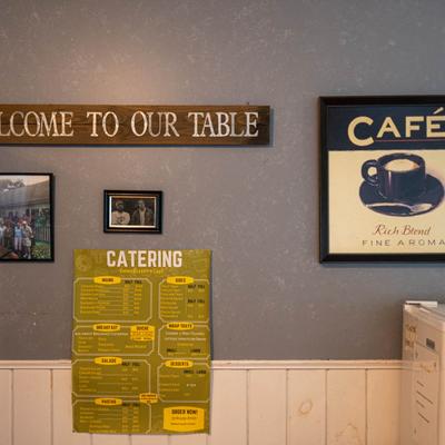 Wall with Welcome to Our Table sign, framed cafe artwork, photos, and a catering menu.