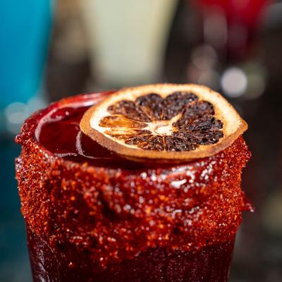 Red Michelada cocktail with a chili rim and dried lemon wheel garnish, close-up.