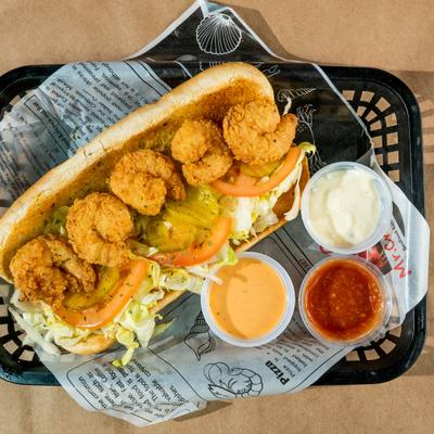 Shrimp po' boy sandwich with lettuce, pickles, and tomatoes accompanied by dipping sauces.