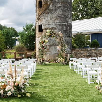 Wedding venue beside a rustic silo, white chairs are arranged on a grassy aisle