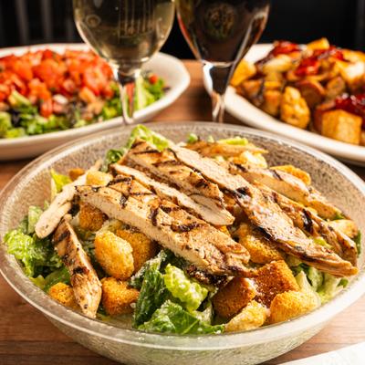 Caesar salad topped with frilled chicken, with glasses of wine and food in the back.