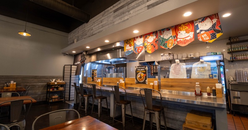 A restaurant interior with a long counter, metallic chairs, and colorful lucky cat banners