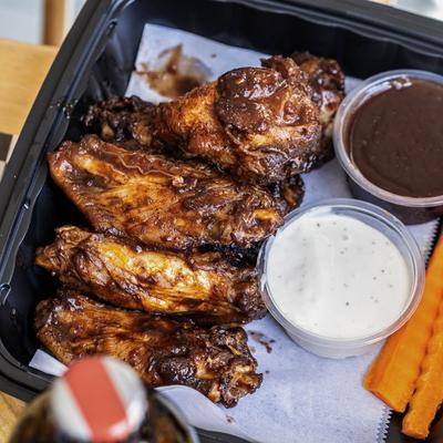 Jerk wings served in a takeout container with cups of sauces and carrots.