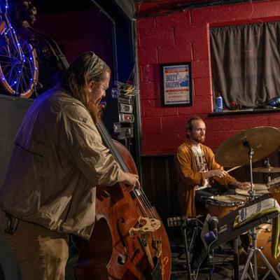 Musicians playing double bass and drums on stage with a red wall background.