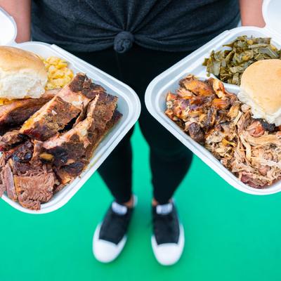 Person holding two barbecue plates with ribs, pulled pork and sides.