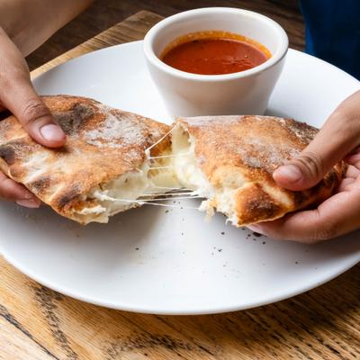 Hands spliting cheese calzone in half with marinara sauce on the side.