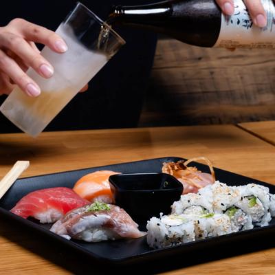 Person pouring drink into a glass, next to a black plate with assorted sushi.