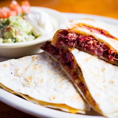 Quesadilla stuffed with corned beef on a plate served with a dish that contains guacamole, sour cream and pico de gallo