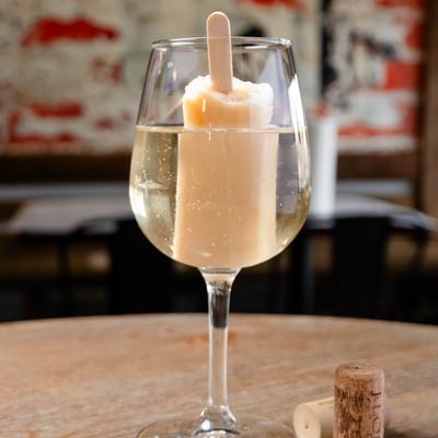 A wine glass filled with sparkling wine and a popsicle inside, rests on a table indoors.