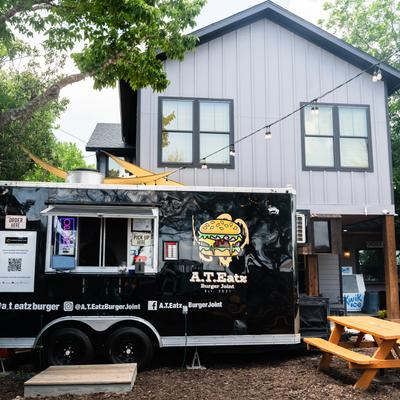 Black food truck with a burger logo under a tree, set near a modern gray house.