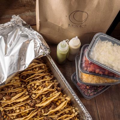 Take-away tray full of tacos with sides, sauces and take-away bag next to it.