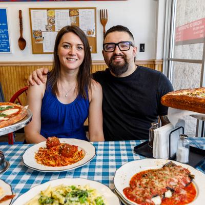 A smiling owners pose at a table with various Italian dishes, including pizza and pasta.