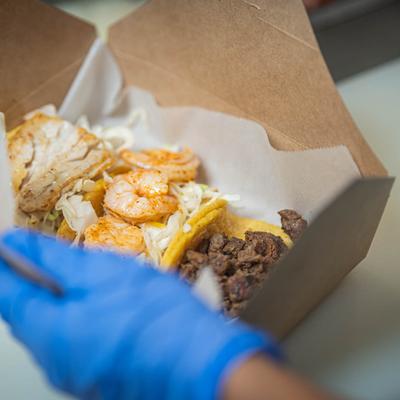 Open takeout box with tacos featuring grilled chicken, shrimp, and beef.