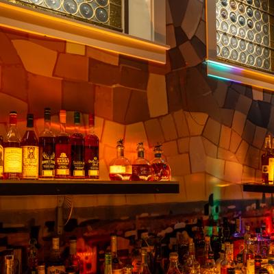 Warmly lit bar shelves displaying assorted liquor bottles against a mosaic patterned wall.