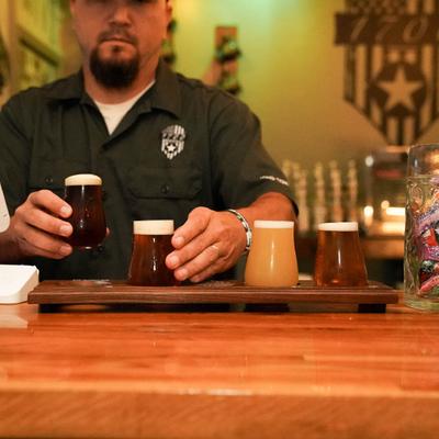 Bartender in a dark shirt serves a flight of four craft beers on a wooden tray.