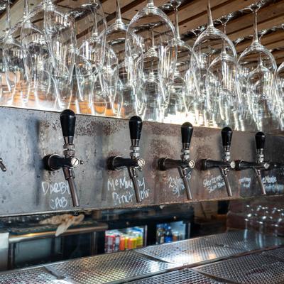 Row of beer taps with handwritten labels beneath hanging wine glasses.