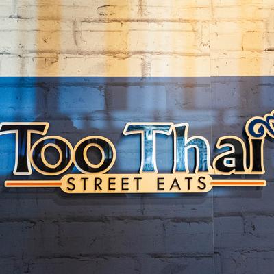 Too Thai Street Eats restaurant sign on a textured brick wall.