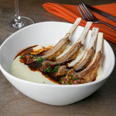 A bowl of creamy cheddar cheese grits topped with four lamb chops and veal demi sauce.
