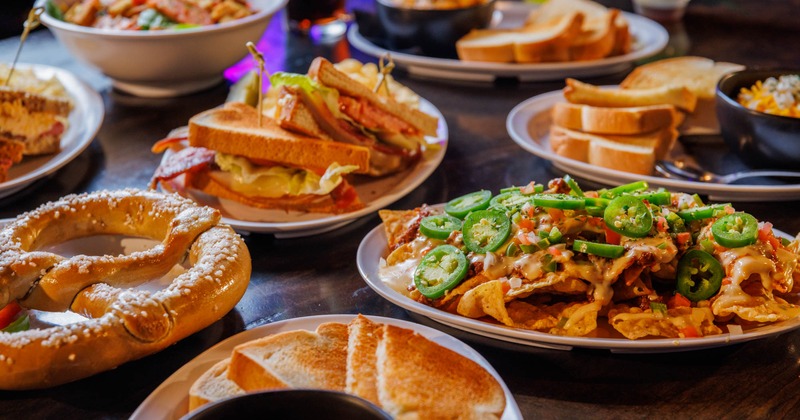 A table full of pub-style food including sandwiches, nachos, toast, and a pretzel