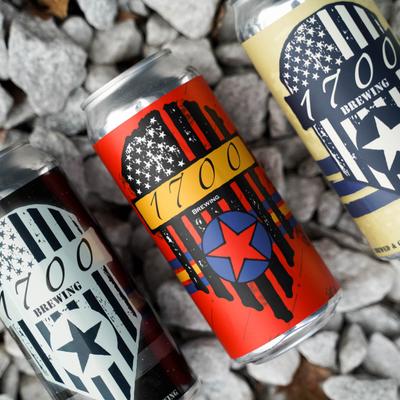 Three cans of 1700 Brewing beer displayed horizontally on gray pebbles.