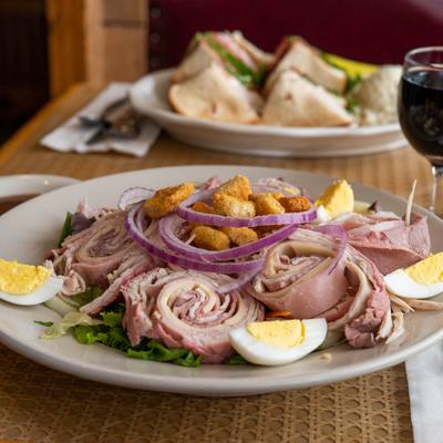 Chef salad with a glass of red wine and club sandwiches in the background.