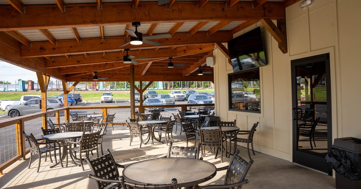 Outdoor patio with a wooden roof, metal tables and chairs, ceiling fans, and a view of a parking lot