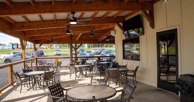 Outdoor patio with a wooden roof, metal tables and chairs, ceiling fans, and a view of a parking lot