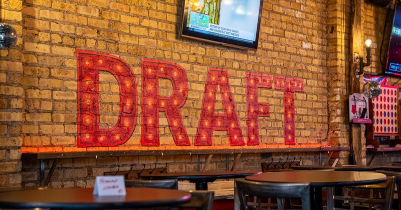 Interior, bar table next to brick wall, sign written on wall, chairs, tv screen above