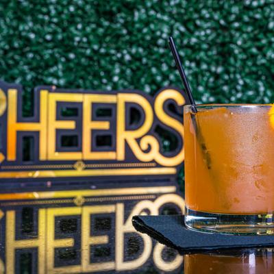 Orange cocktail drink served on a table with cheers sign and faux greenery wall.