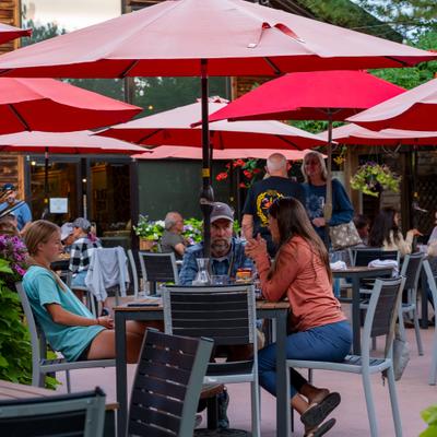 Guests dining outdoors under red patio umbrellas at a busy restaurant.