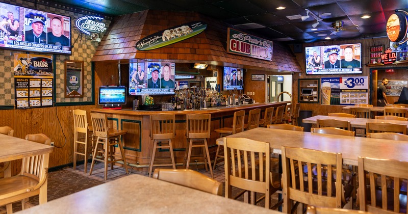 Tables and chairs arranged in front of a bar with multiple TV screens and wall decorations
